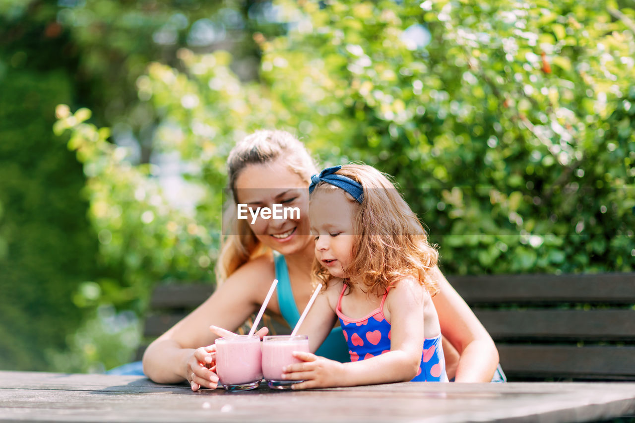 A beautiful girl and her little daughter drink a refreshing smoothie while sitting on a bench 