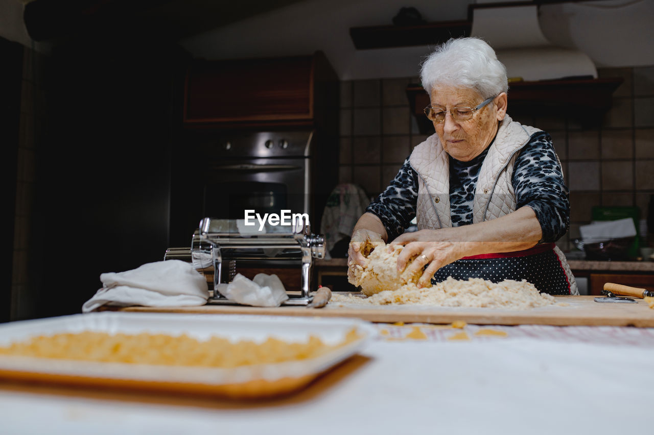 Senior female standing at table in cozy kitchen and kneading raw dough while preparing pastry for cooking domestic italian tortellini