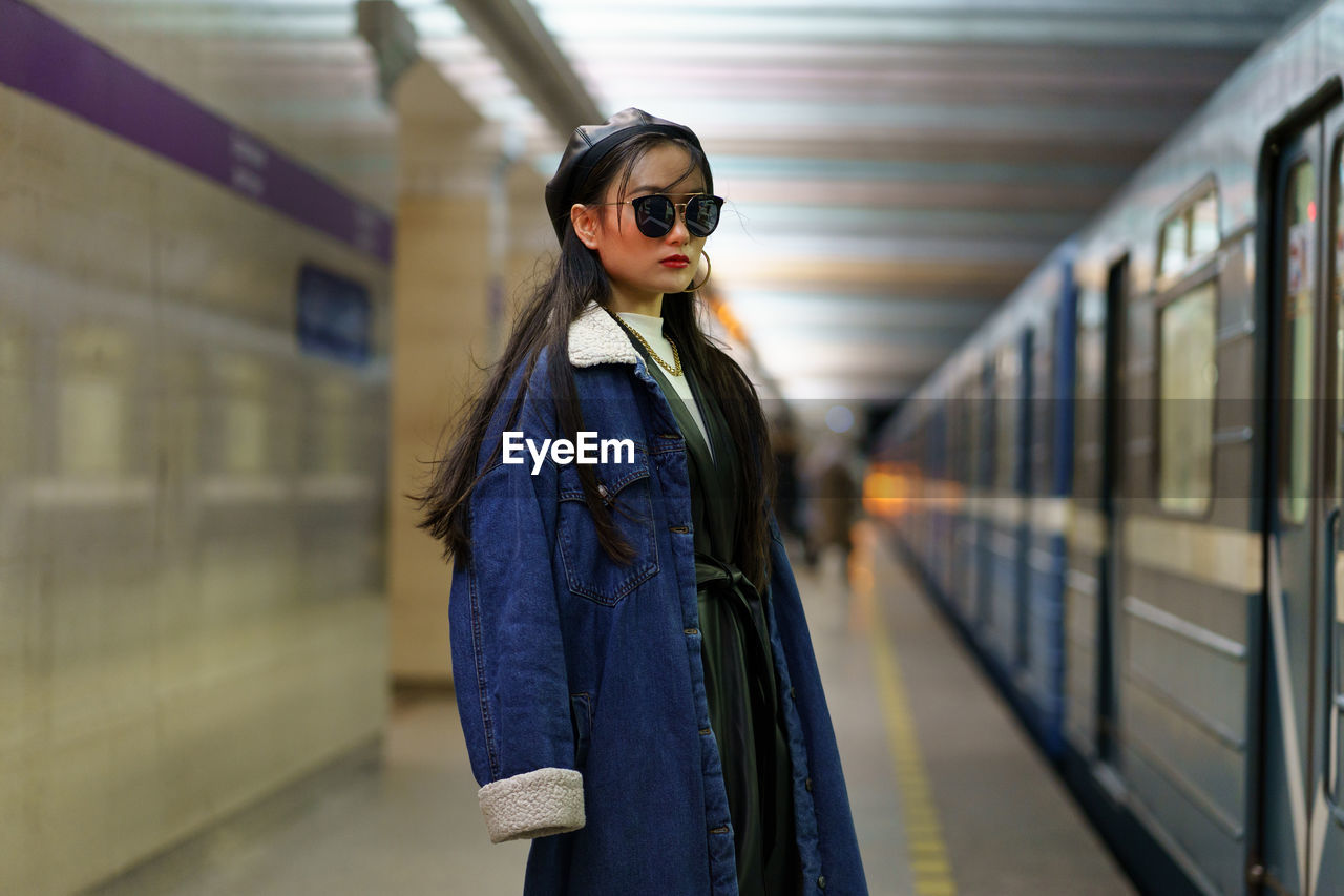 Stylish asian girl in glasses and street fashion outfit at underground platform with arriving train