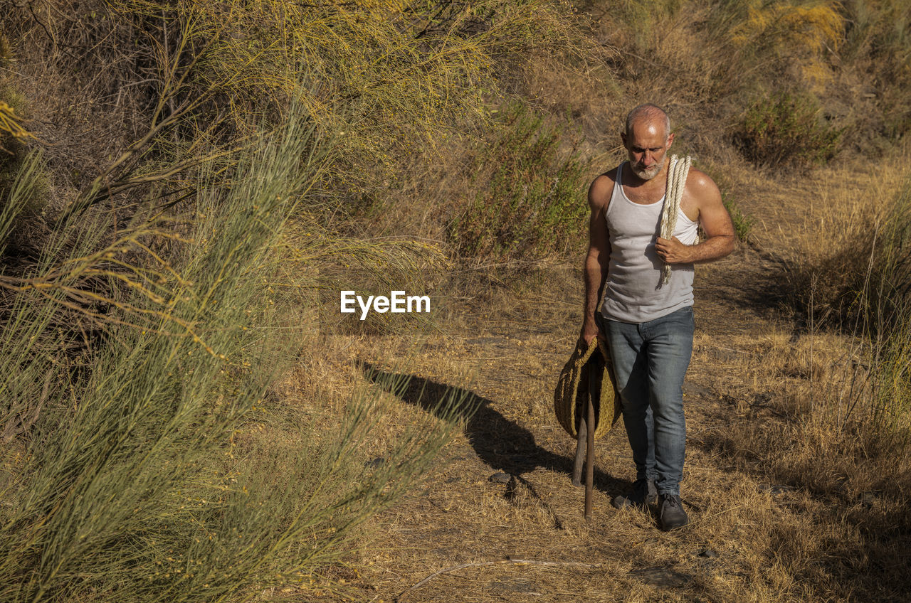 Full length portrait of man in white tank top and jeans walking on path in fields in summer