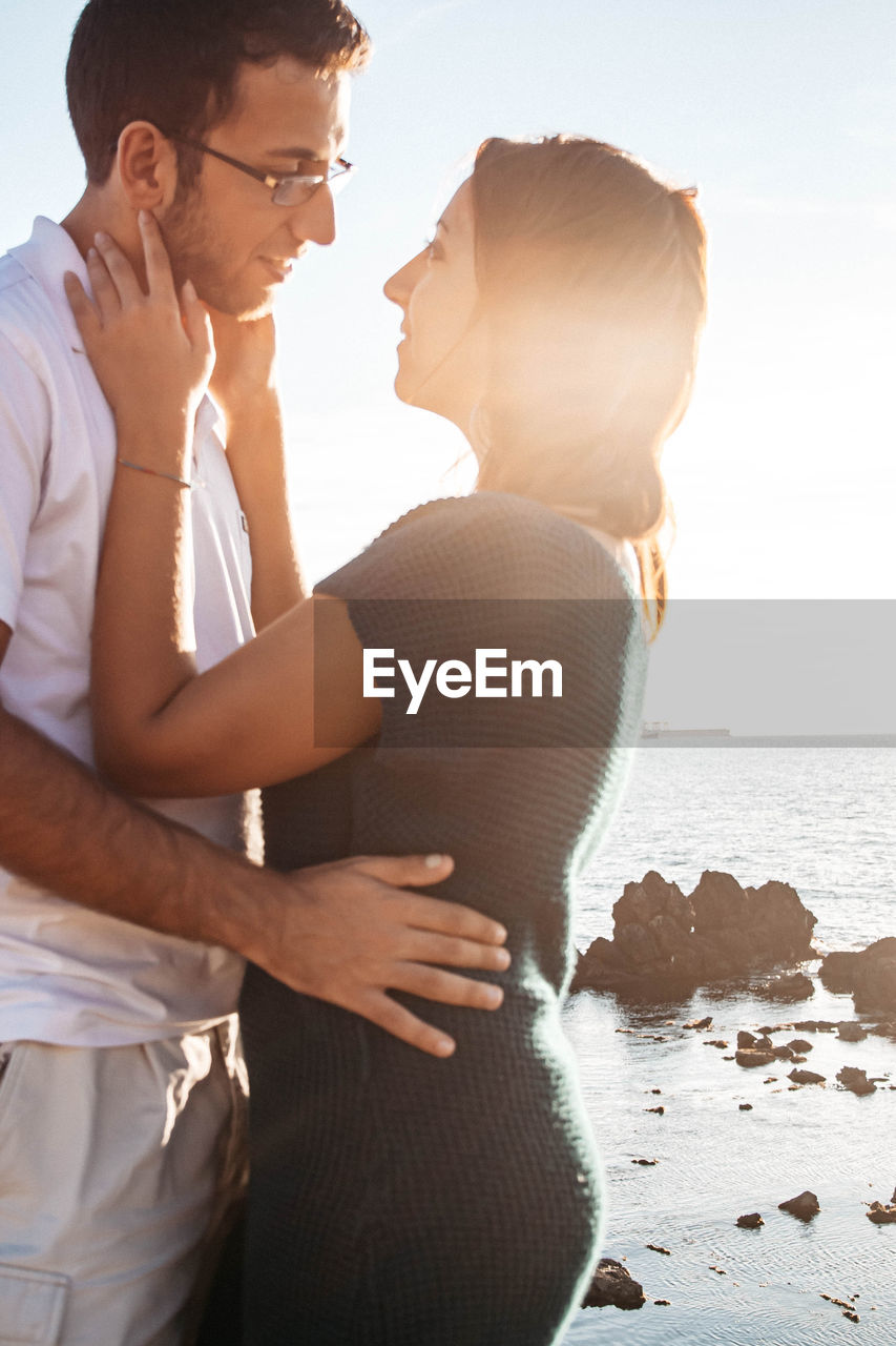 Photo of a couple looking at each other romantically standing in front of a sunset over the sea