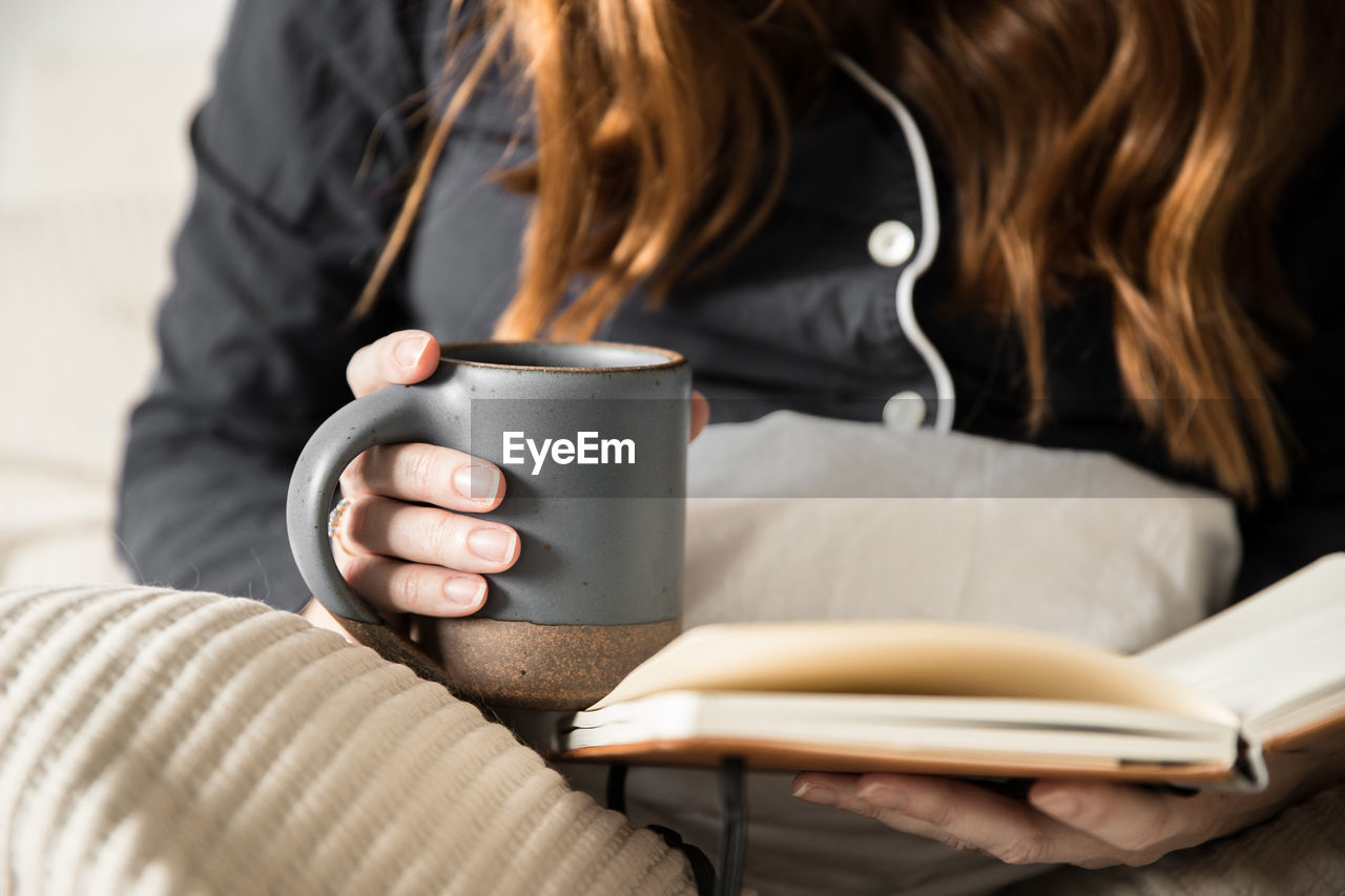 Midsection of woman holding mug while reading book on bed