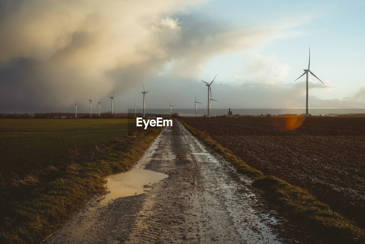 Road amidst field against cloudy sky during sunset with wind turbine 