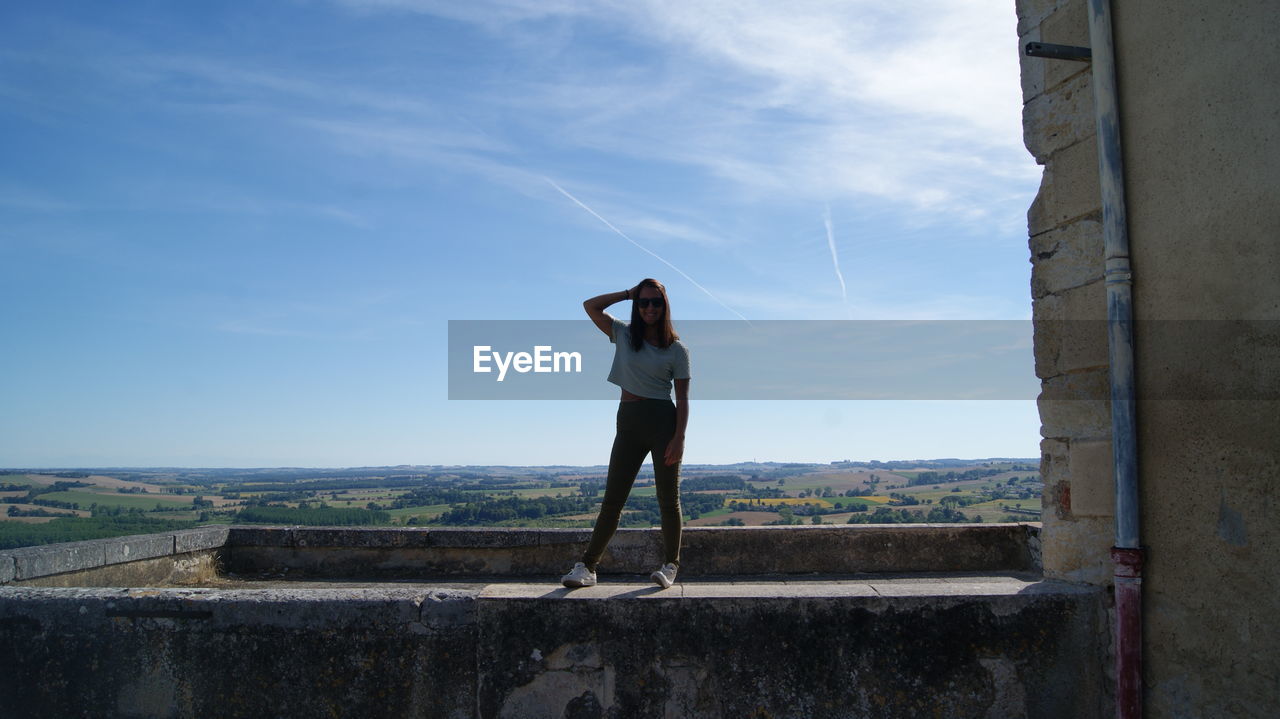 Full length of carefree woman standing on retaining wall against sky