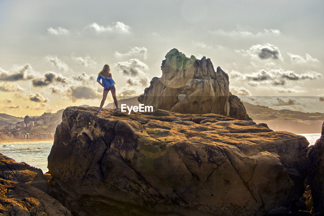 Rear view of mid adult woman standing on rock against sky during sunset