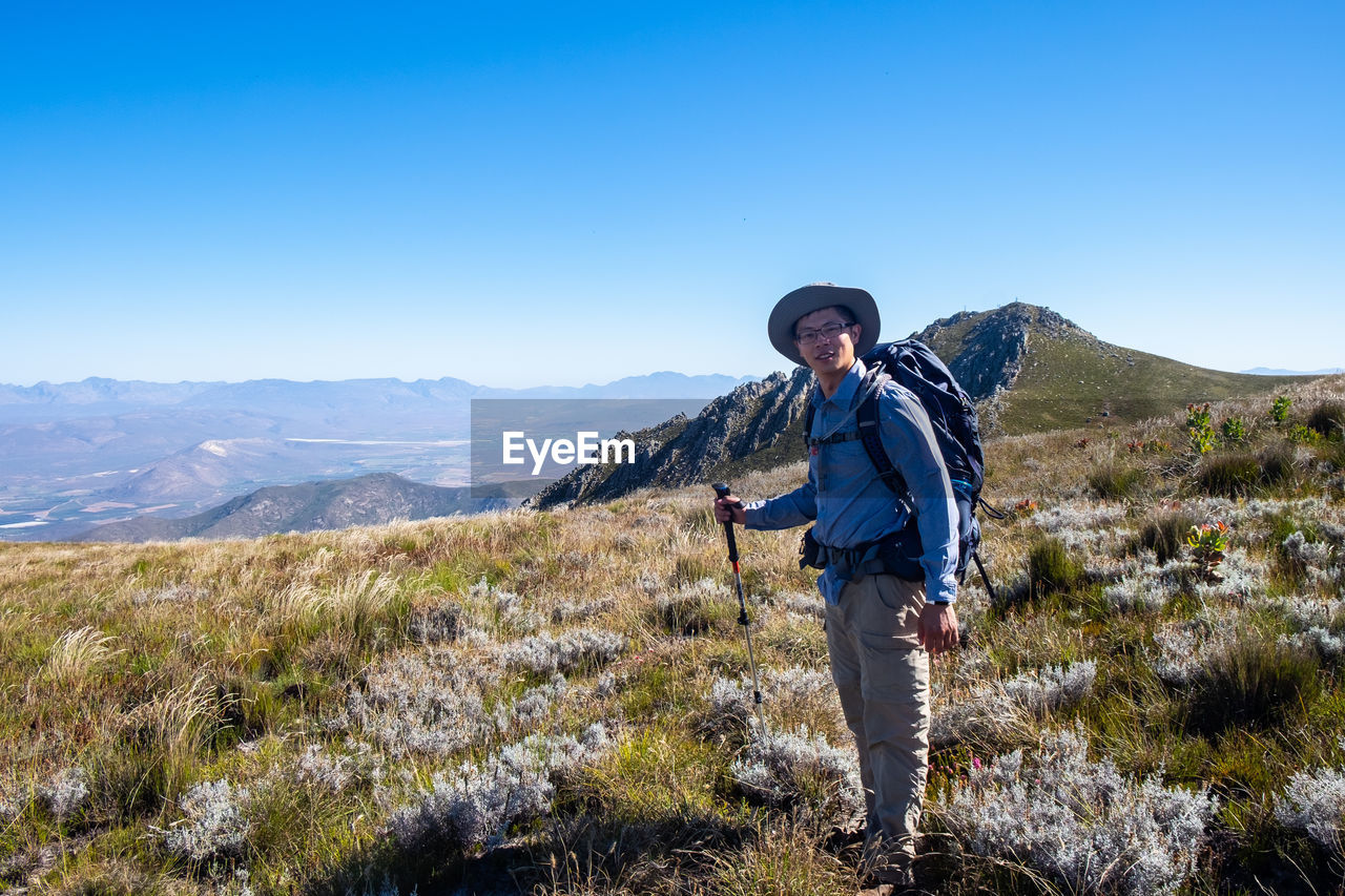 Man with backbag and trekking pole standing in wild flower field with mountain as background 