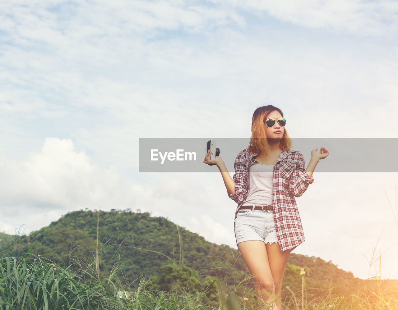 PORTRAIT OF YOUNG WOMAN HOLDING SMART PHONE WHILE STANDING ON MOUNTAIN AGAINST SKY