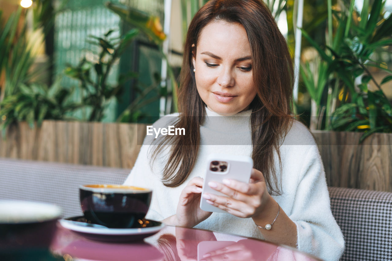 Attractive young brunette woman in white casual dress with cup of coffee using mobile phone in cafe