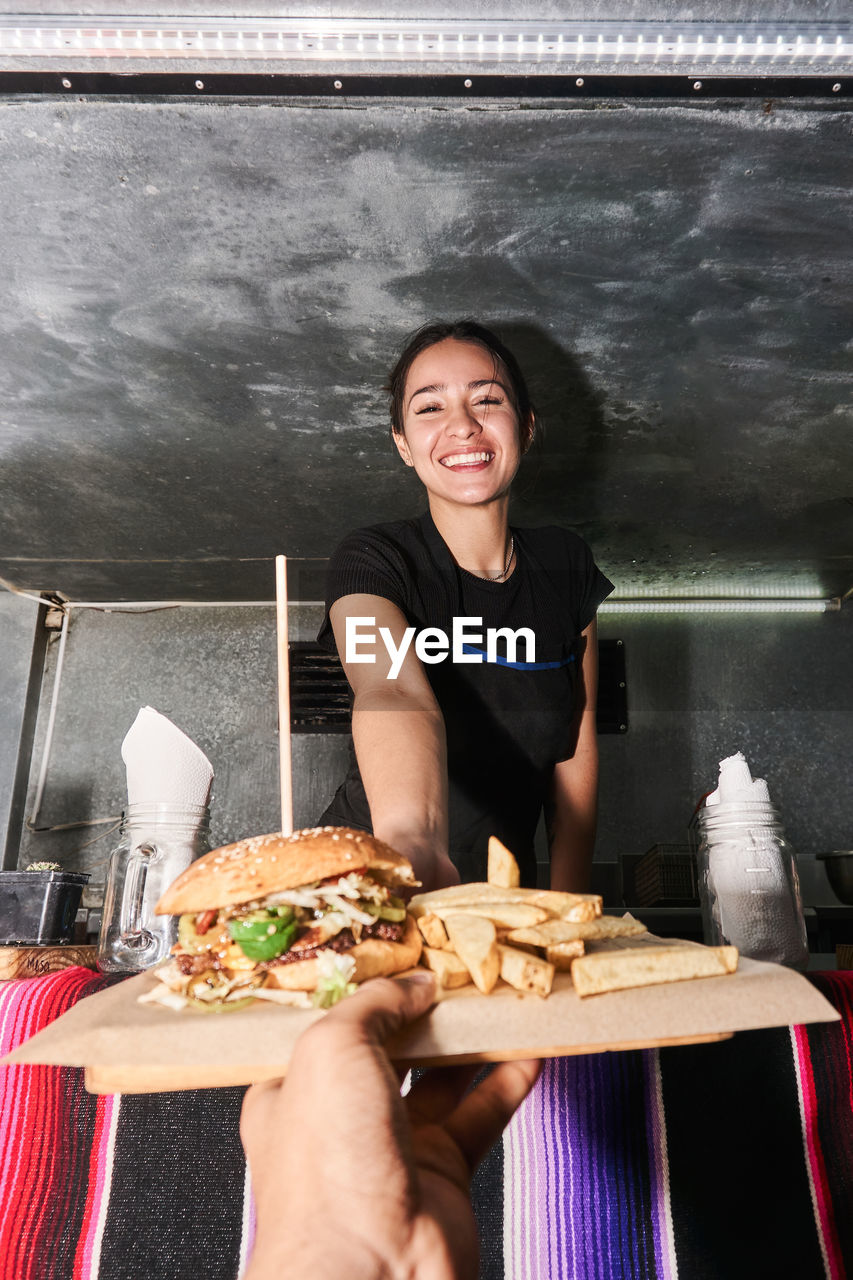 Hand of faceless unrecognizable buyer getting order with fast food from happy ethnic saleswoman at truck counter