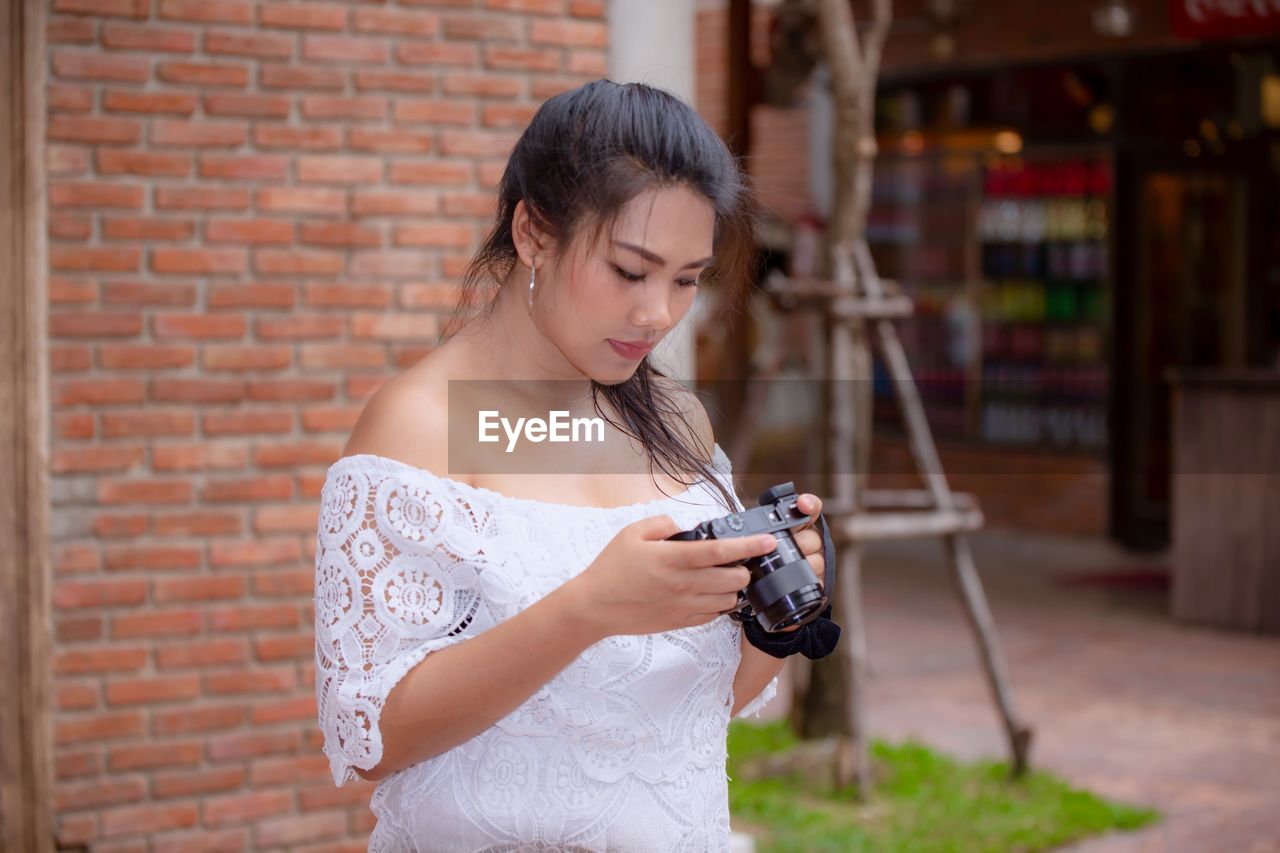 Young woman holding camera while standing by brick wall