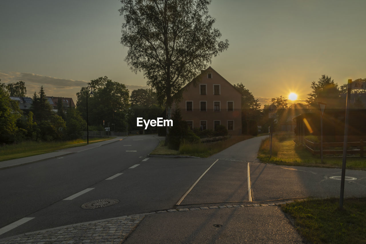 morning, tree, architecture, sky, plant, road, built structure, building exterior, nature, transportation, city, sunlight, building, street, light, no people, house, lane, sign, residential area, residential district, sun, dusk, outdoors, symbol, lens flare, road marking, suburb, cloud