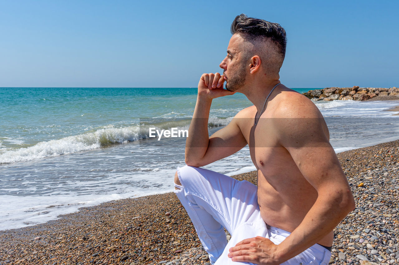 REAR VIEW OF MAN SITTING ON BEACH AGAINST SKY