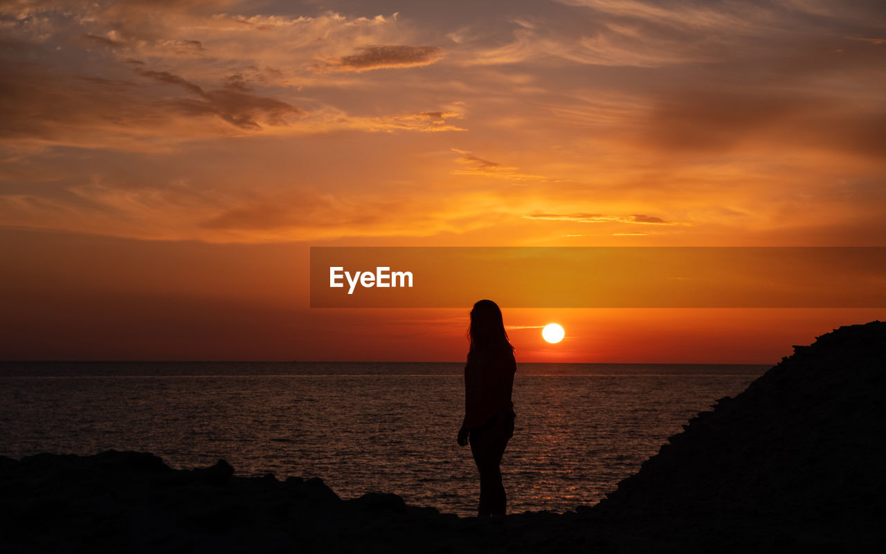 Silhouette woman standing at beach against sky during sunset