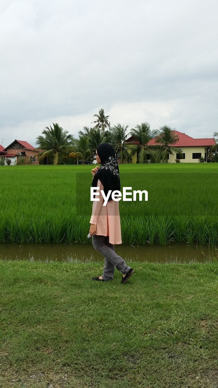 Side view of teenage girl walking at rice paddy against cloudy sky