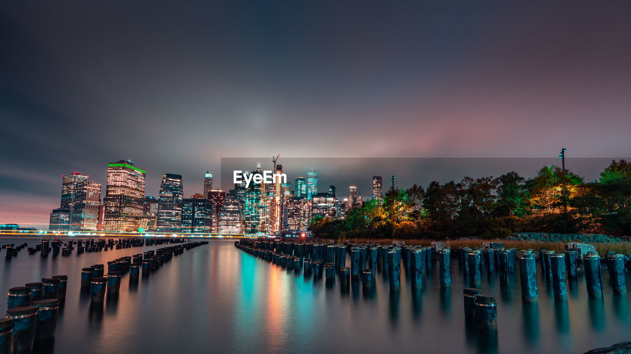 Illuminated modern buildings by river against sky at night
