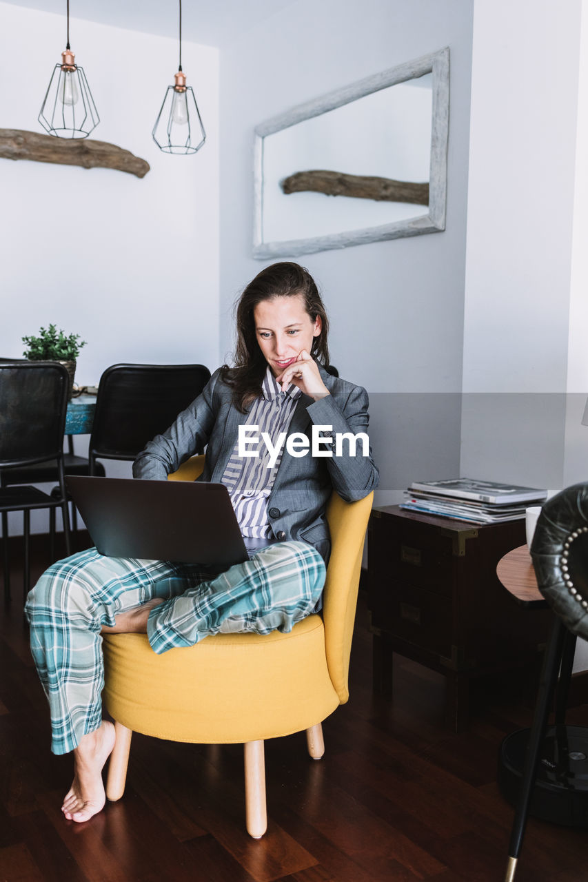 Full body of young female remote employee wearing formal blazer and homey pajama pants sitting on chair and working on laptop at home