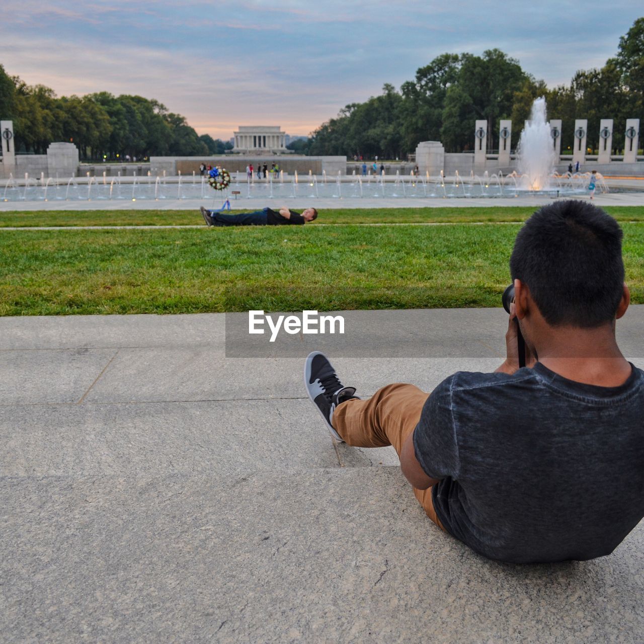 Rear view of man photographing friend lying by fountain against sky