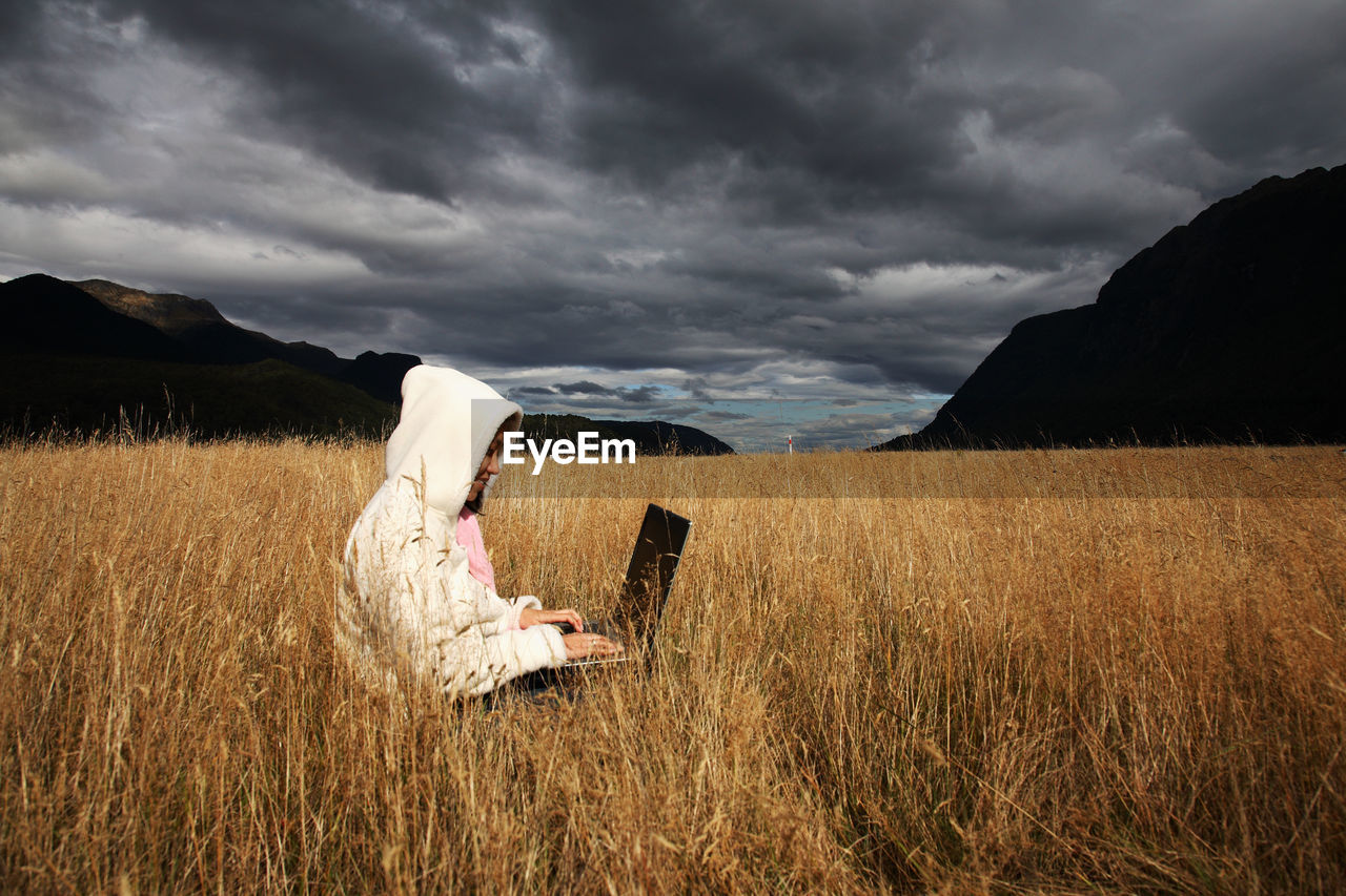 Mid adult woman using laptop while sitting on grassy field against cloudy sky