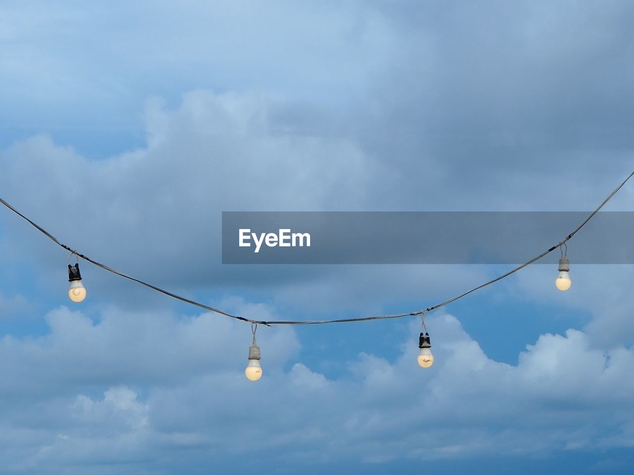 Low angle view of light bulbs hanging against cloudy sky