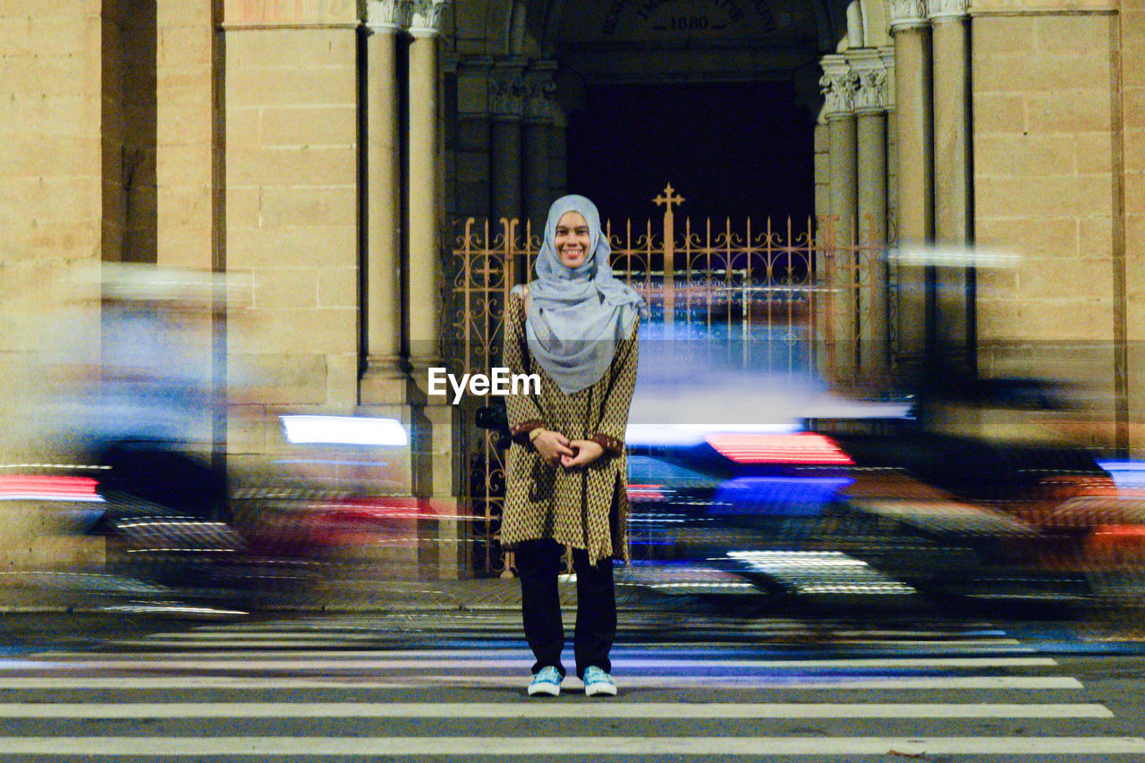 Full length portrait of smiling woman in hijab standing on city street with blur traffic in background at night