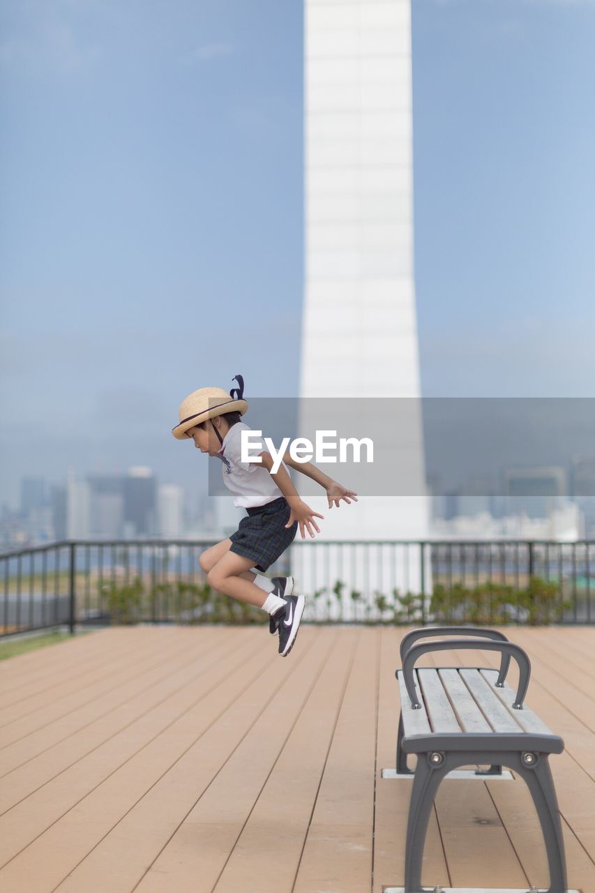 Side view of boy wearing hat while jumping on footpath against sky