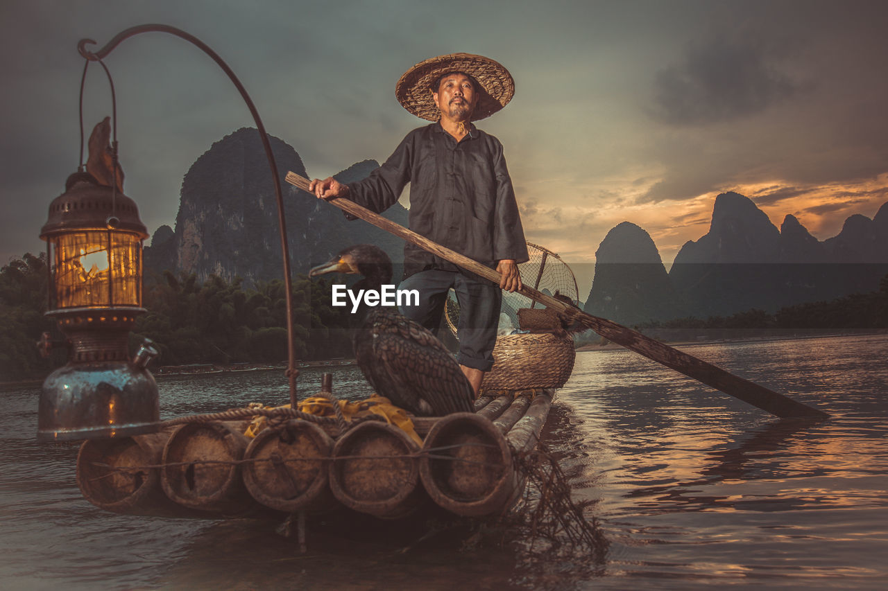 Portrait of traditional fisherman by cormorants on wooden raft over river at sunset