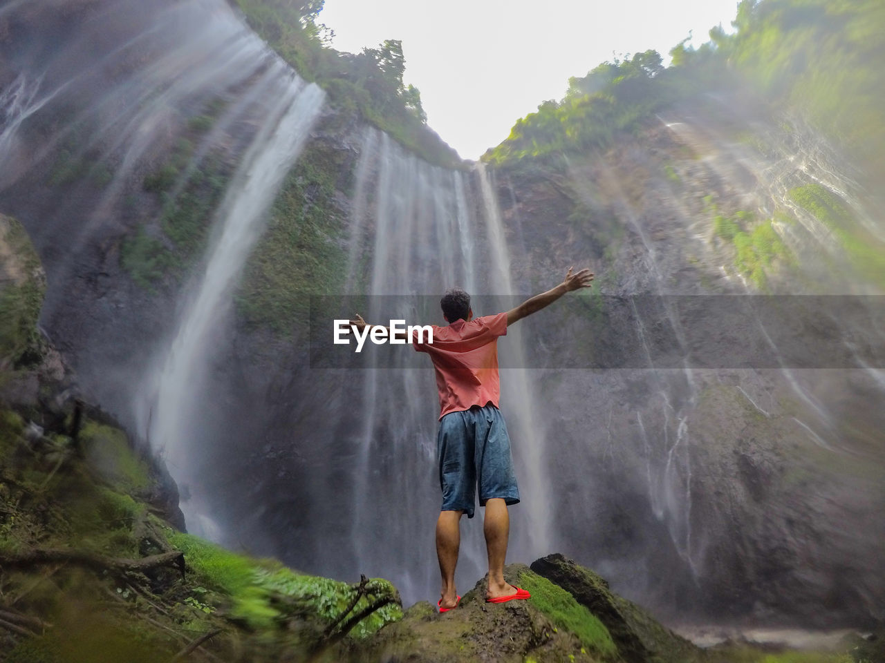 Rear view of man outstretching arms in front of waterfall