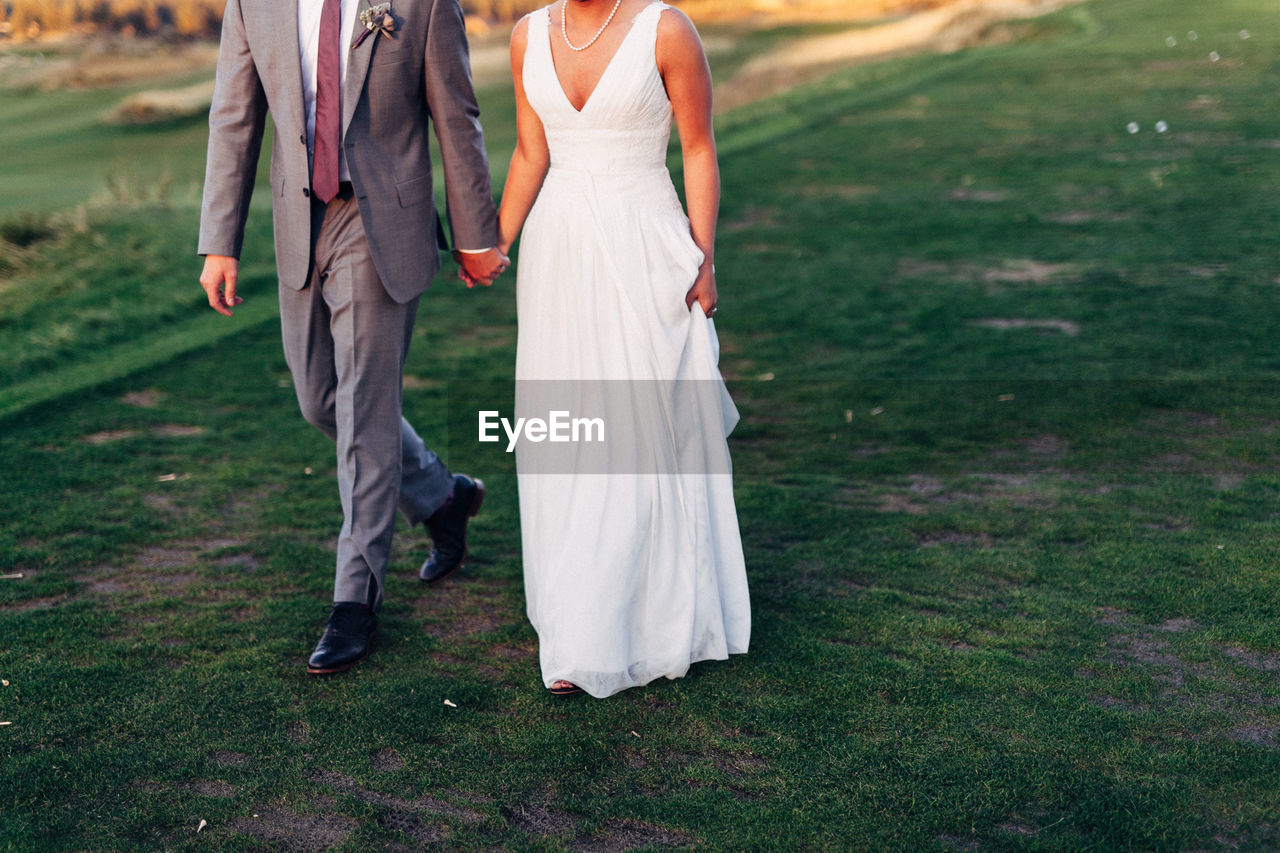 Low section of wedding couple holding hands while walking on field