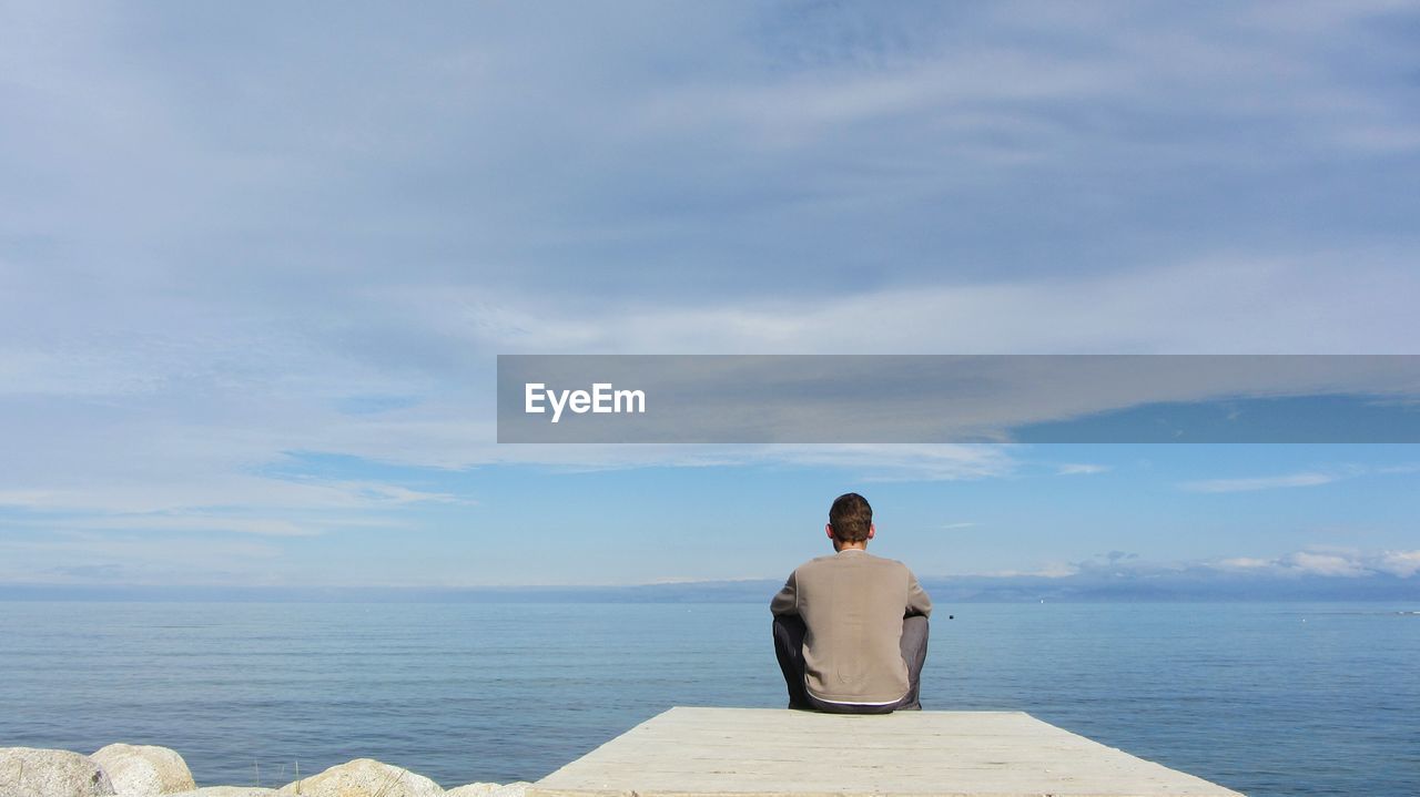 Rear view of man sitting on pier in sea against cloudy sky