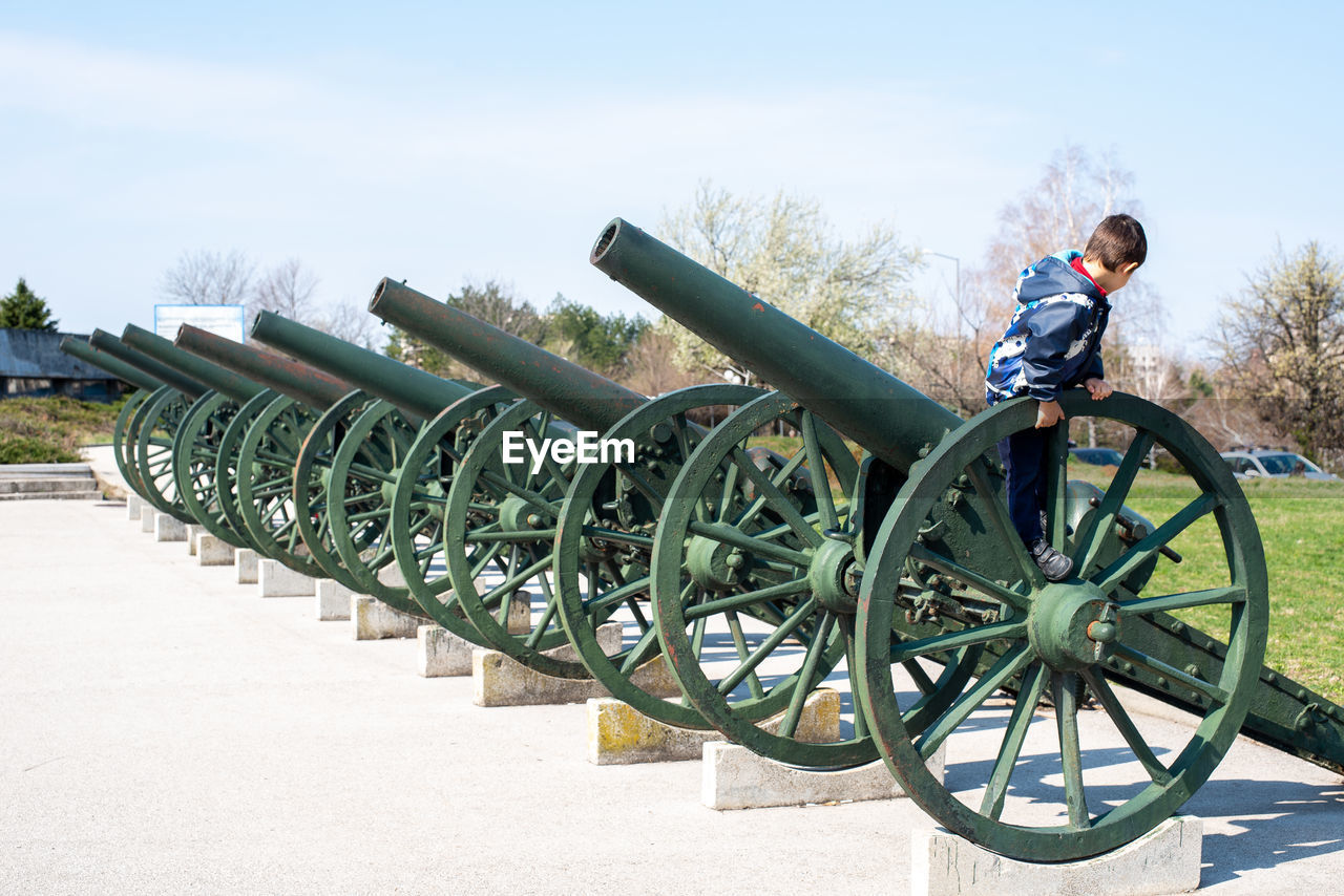 Side view of boy standing on gun barrel against sky