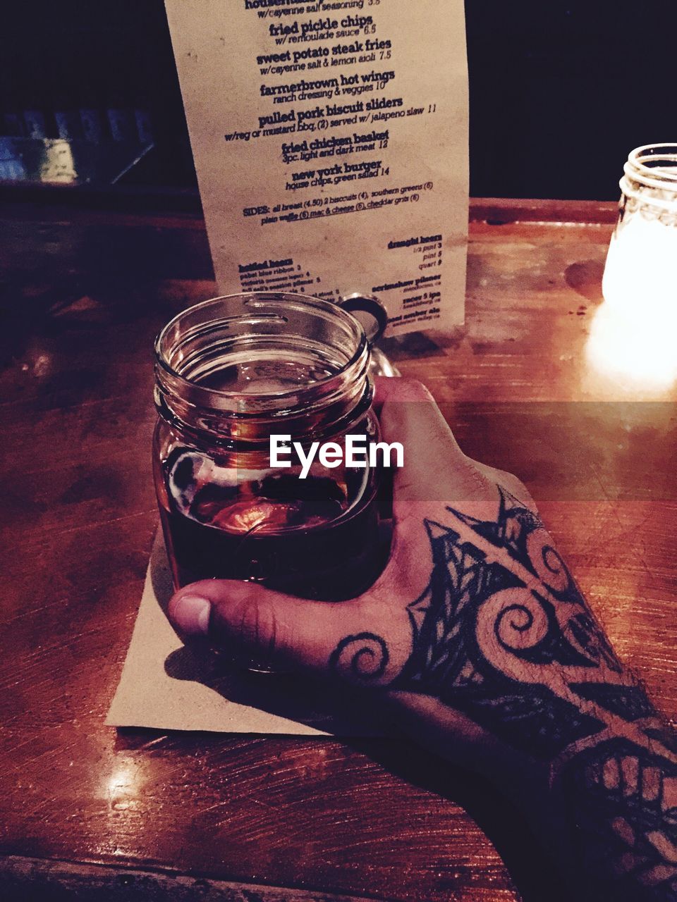 Cropped image of tattooed hand holding glass jar of drink at table in restaurant