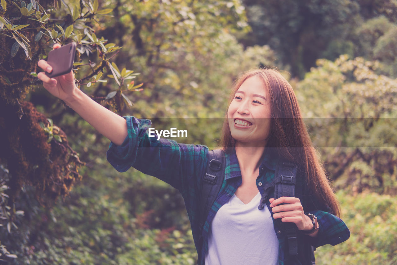 Smiling woman taking selfie with smart phone while standing against trees in forest