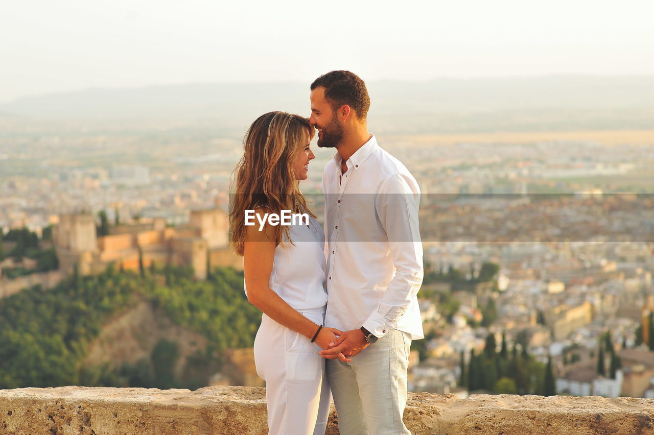 Side view of young romantic couple standing by retaining wall against city