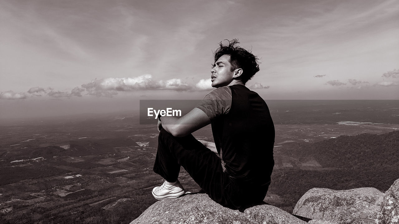 Side view of young man sitting on rock looking at landscape