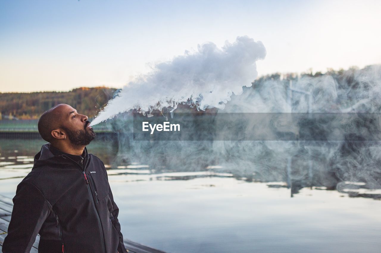 Side view of mid adult man emitting smoke while standing by lake against sky