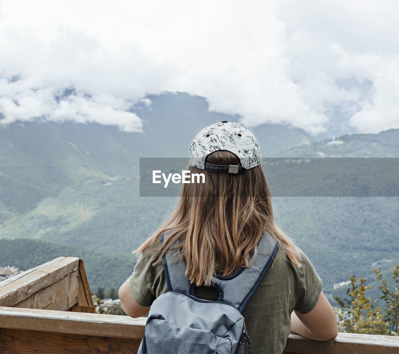 Rear view of young blonde woman in cap looking at view of caucasian mountains and cloudy sky in hike