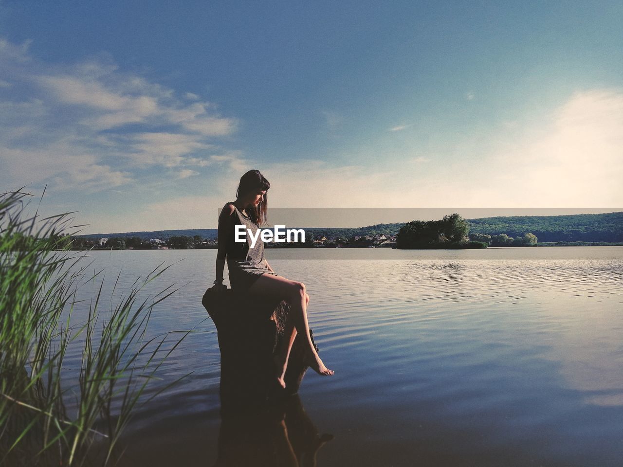 Woman sitting on wooden post over lake during sunset