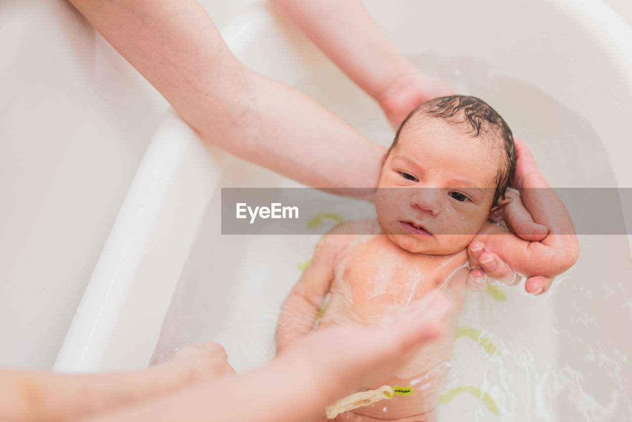 From above unrecognizable parent washing crying newborn baby in warm water in basin at home