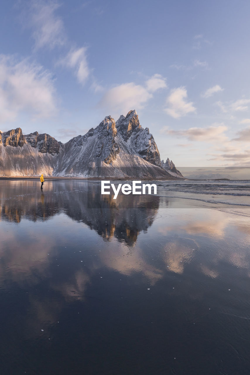 Anonymous tourist standing on wet shore near calm sea and rough mountains during majestic colorful sunset