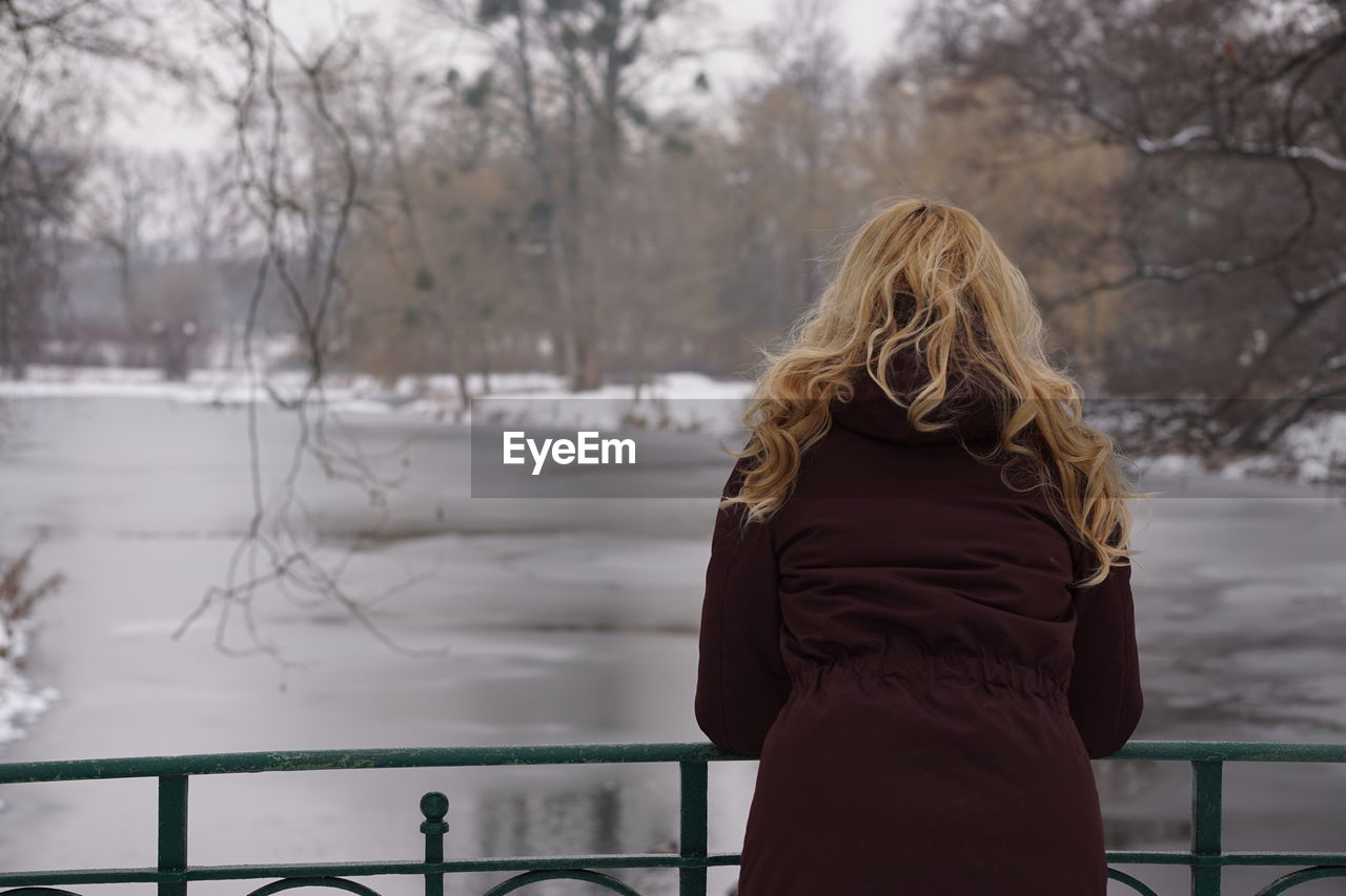 Rear view of woman leaning on railing at footbridge over frozen lake during winter