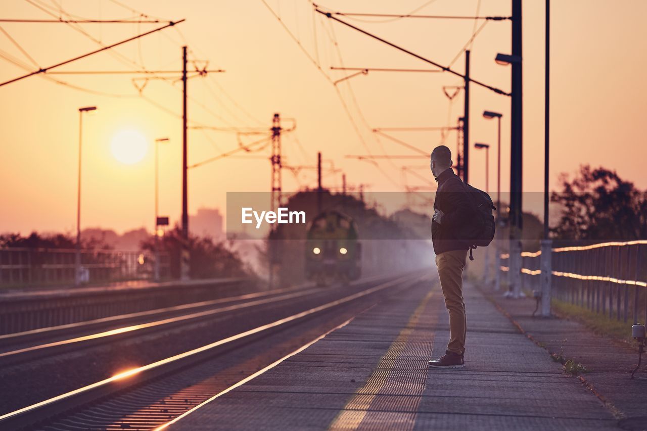 Side view of man standing at railroad station against sky during sunset