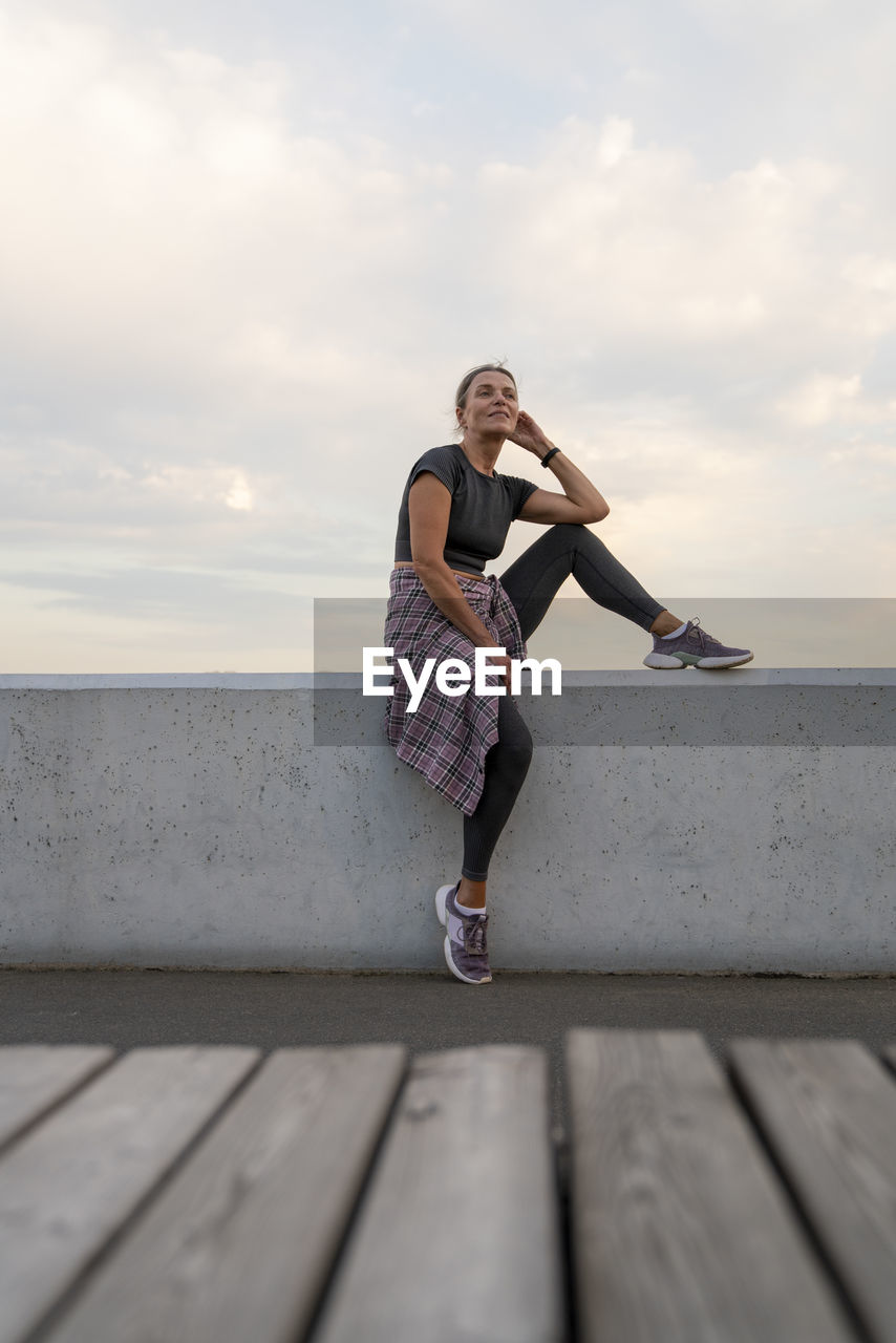 Mature woman wearing sports clothing sitting on wall in front of sky