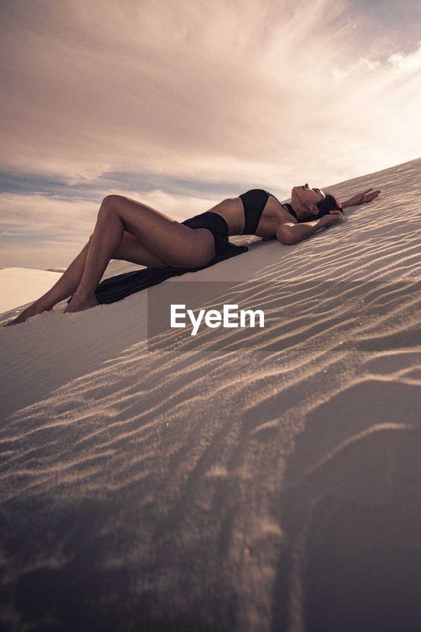 Side view of seductive woman lying on desert against sky