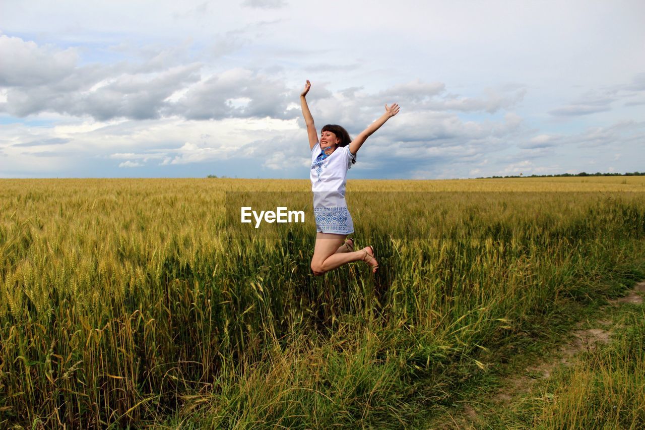 Cheerful young woman with arms raised jumping on field against cloudy sky