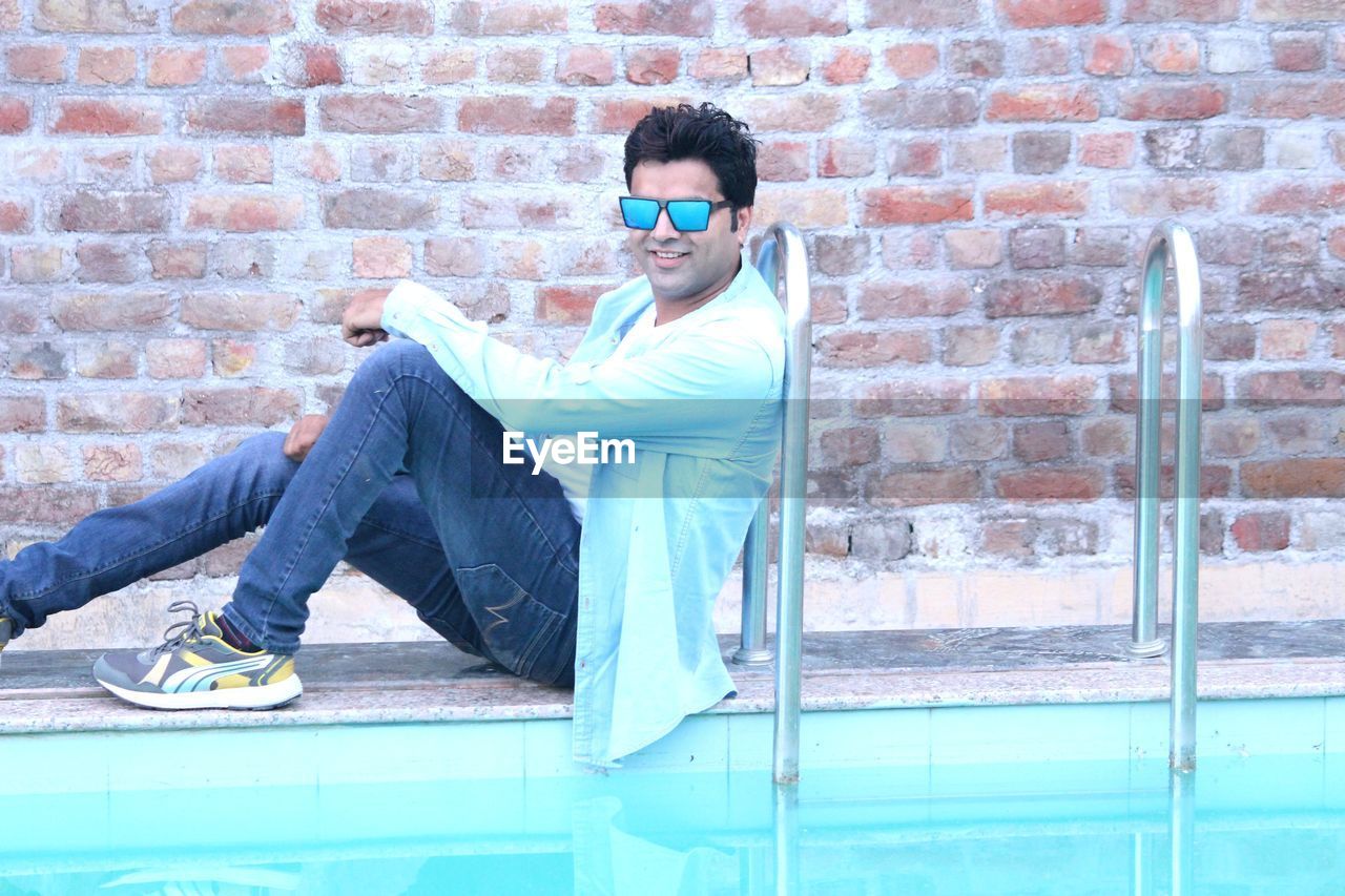 FULL LENGTH PORTRAIT OF YOUNG MAN SITTING AGAINST BRICK WALL