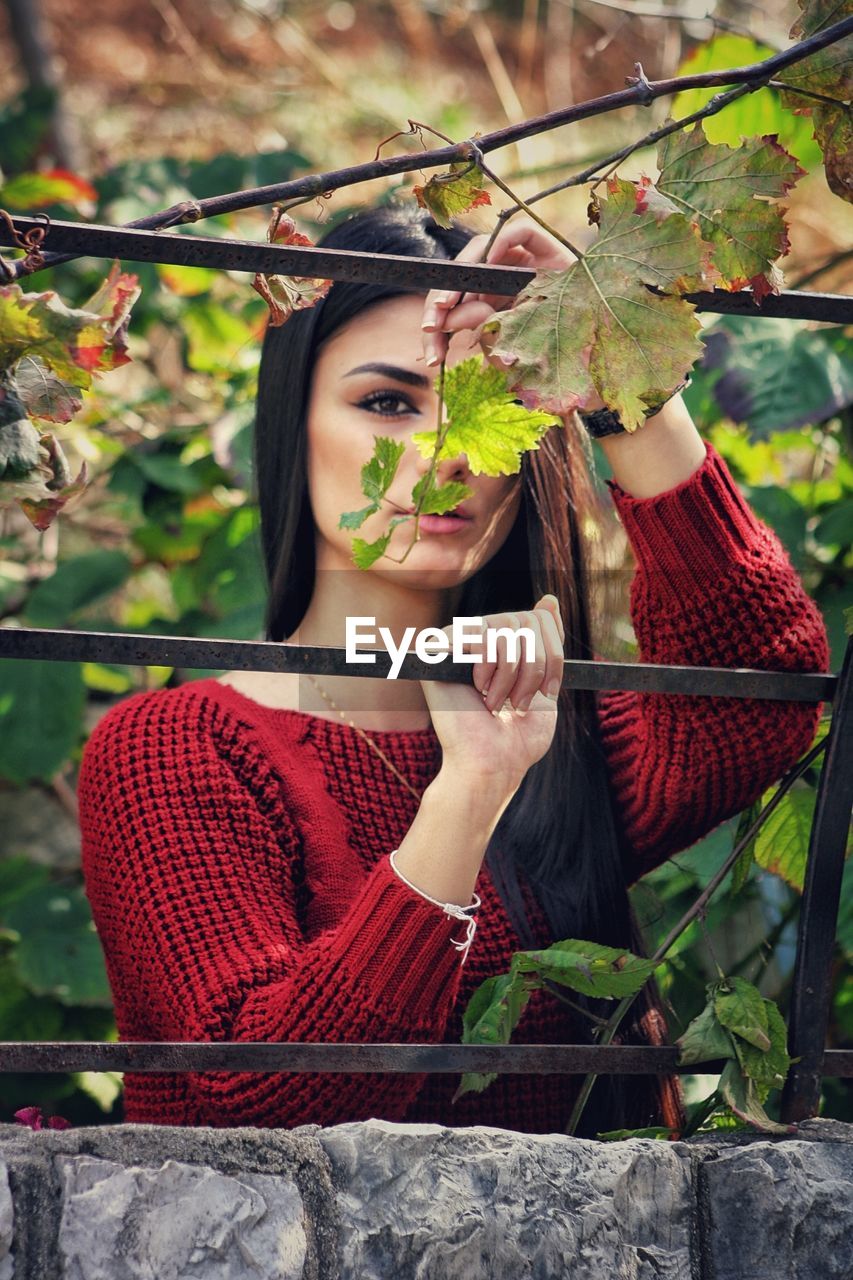 Portrait of young woman seen through fence standing against plants