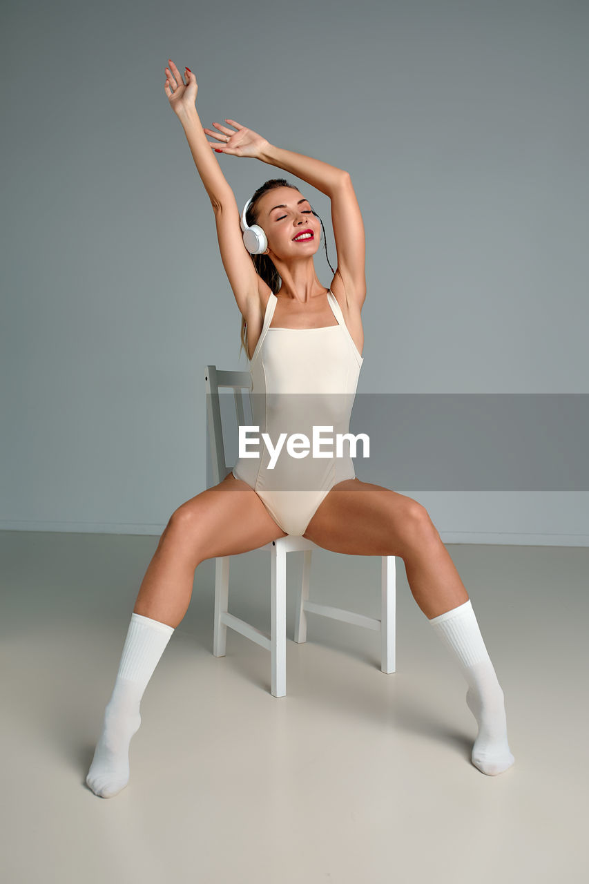 women, one person, studio shot, adult, full length, indoors, young adult, gray background, sports, gray, limb, human limb, arm, portrait, arms raised, lifestyles, sitting, clothing, smiling, happiness, human leg, female, wellbeing, colored background, balance, posture, ballet, emotion, vitality, flexibility, dancing, exercising, looking, leotard, front view, performing arts, stretching, team sport, seat, cheerful, ballet dancer, modern dance, elegance