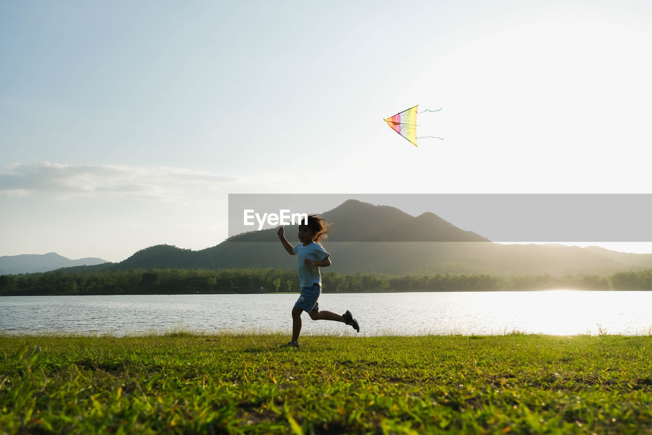 Rear view of man with arms outstretched standing by lake against sky during sunset