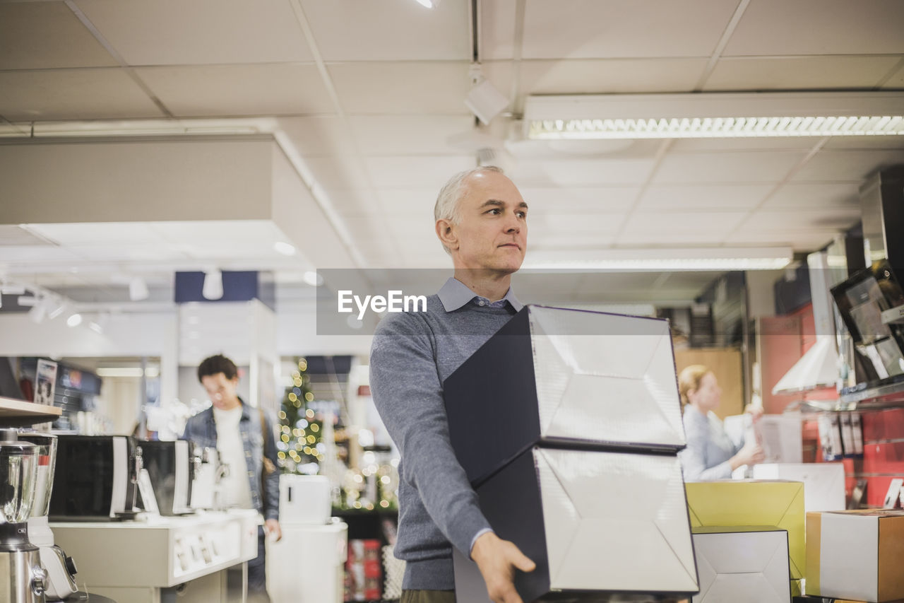 Mature salesman with boxes looking away while walking in electronics store