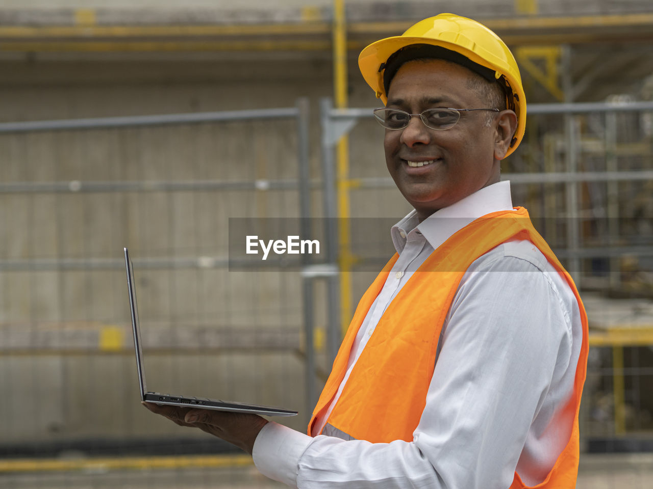 Smiling indian engineer wearing a safety helmet and jacket holding a laptop computer